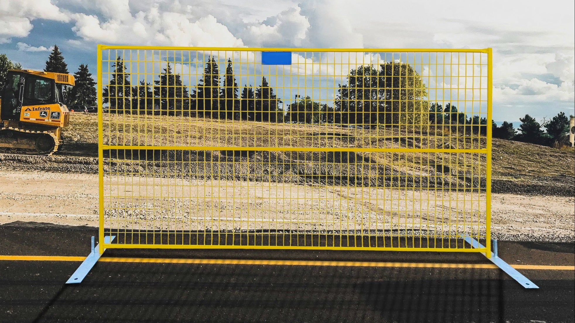 A bright yellow temporary metal mesh fence with blue ground anchors, set up on an asphalt surface at a construction site under a cloudy sky.